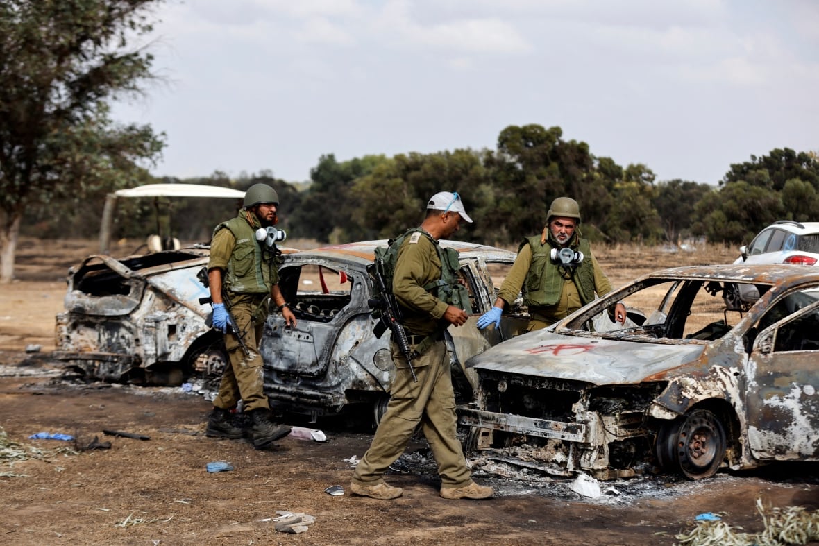 Three soldiers in green uniforms and helmets walk around the remains of burnt cars.