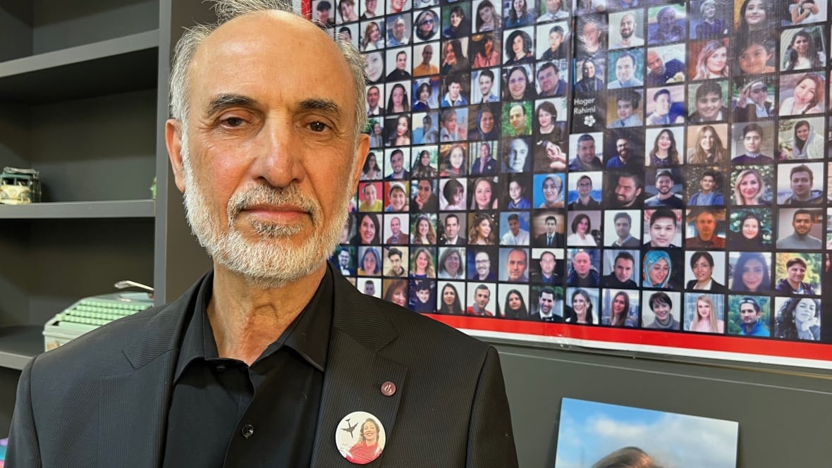 A balding man in a black suit with a button featuring a photo of a young woman on his lapel stands in front of a poster of dozens of photos of people on the wall.