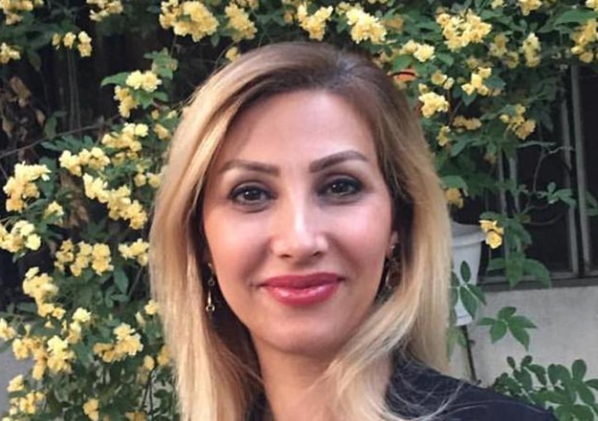 A woman with blond hair stands in front of a bush with yellow flowers, smiling at the camera.