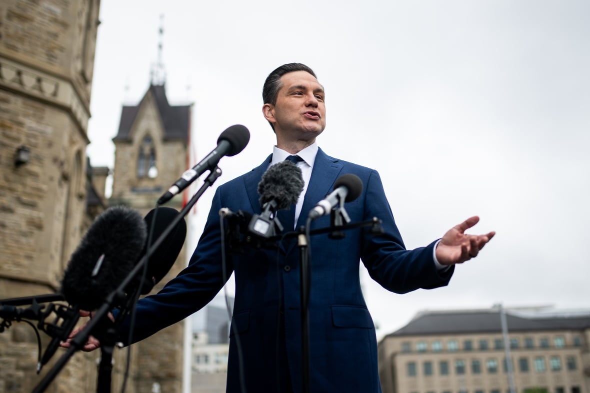 A man in a suit gestures while speaking, outside, in front of several microphones.