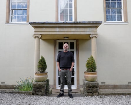 A man in black sweater and jeans stands between pillars in the doorway of a tall, pale stone building