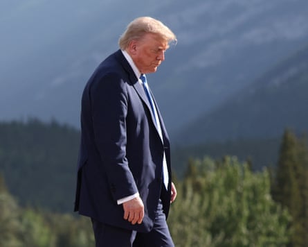 Donald Trump in side profile standing on a mountainside with trees in the background; he wears a dark blue suit and is looking downward