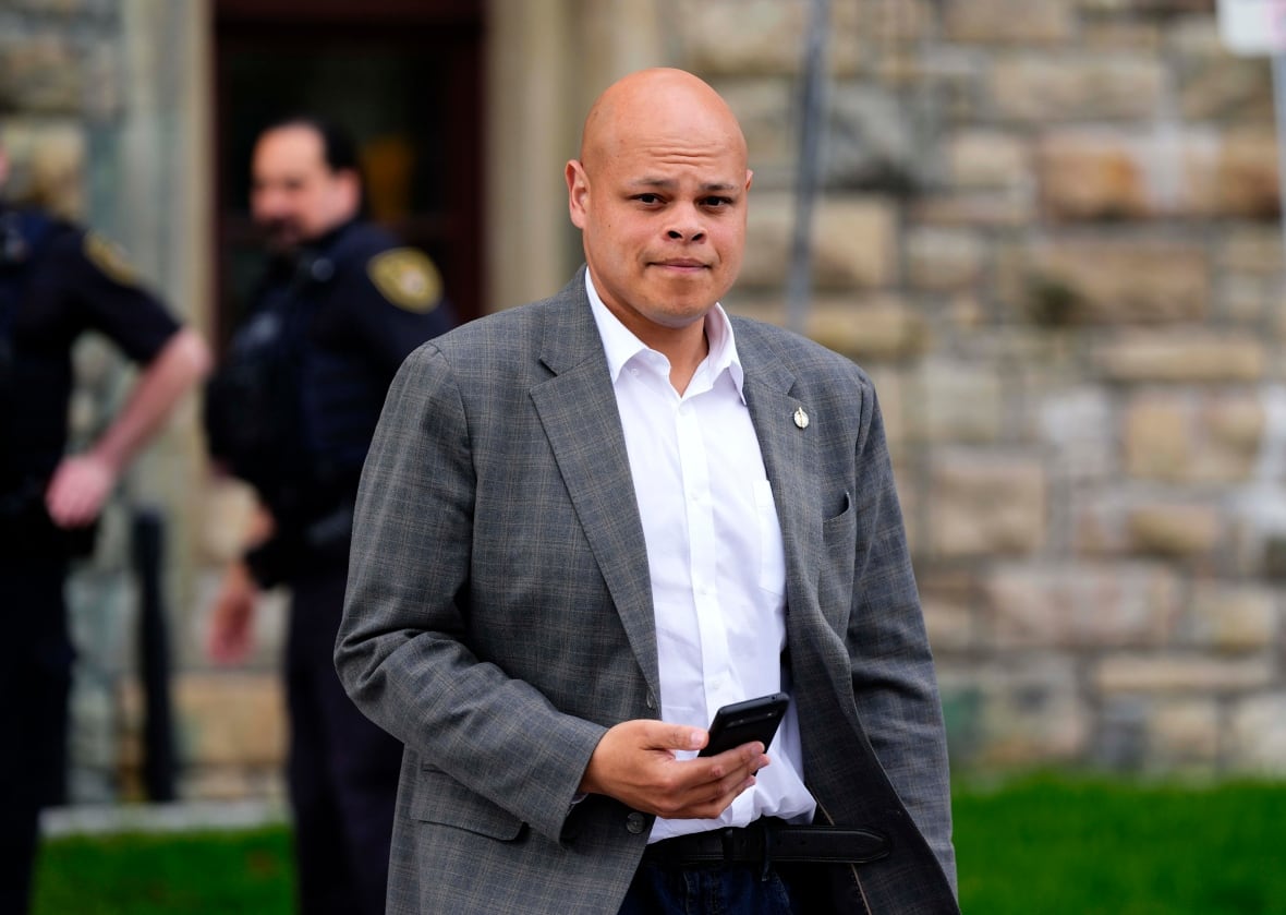 Conservative MP Jamil Jivani is shown walking outside wearing a blazer with no tie. He looks at the camera as he walks while holding a mobile phone in his right hand. 