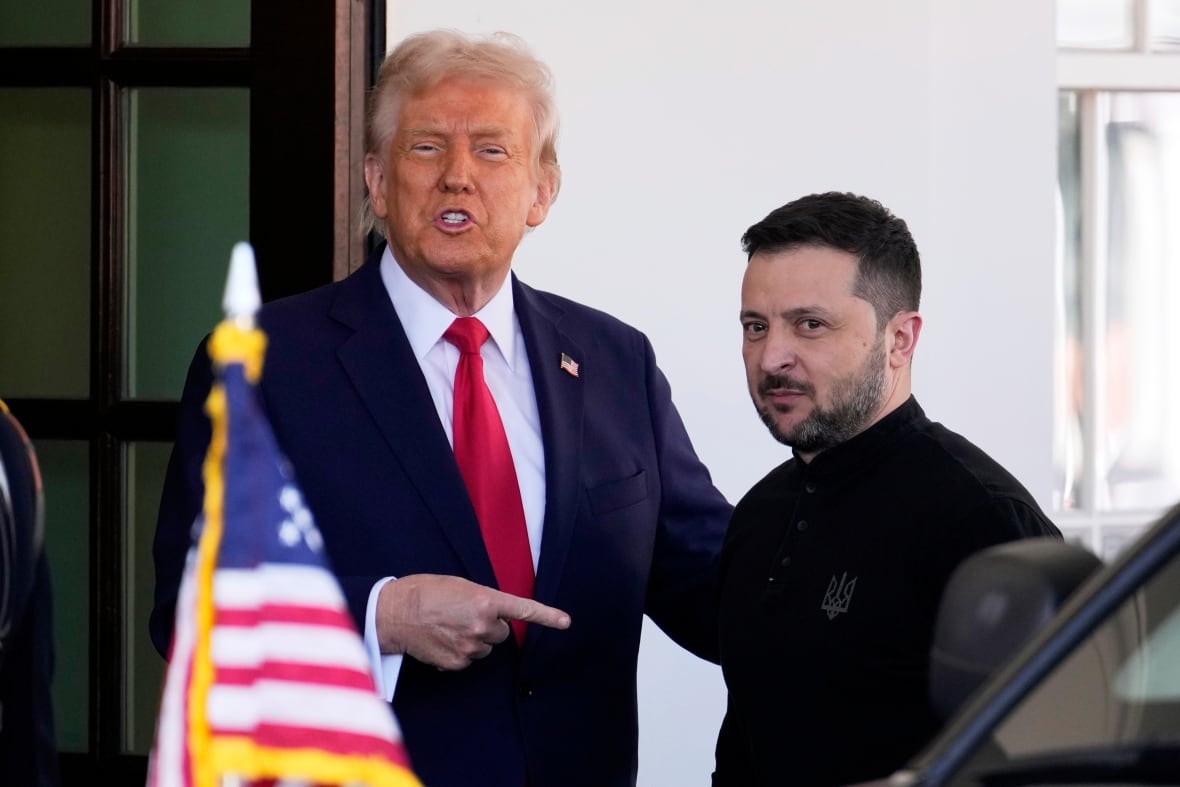 A man in a suit points to another man. They are side by side and near an American flag.