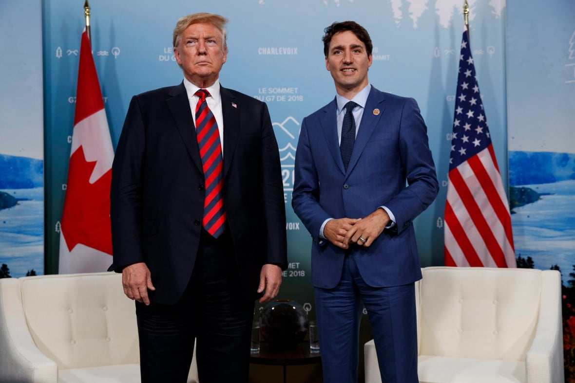 Two men in suits stand together by Canadian and American flags.
