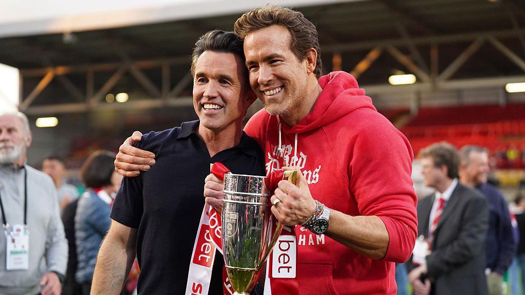 Ryan Reynolds and Rob McElhenney celebrate promotion with a trophy on the pitch