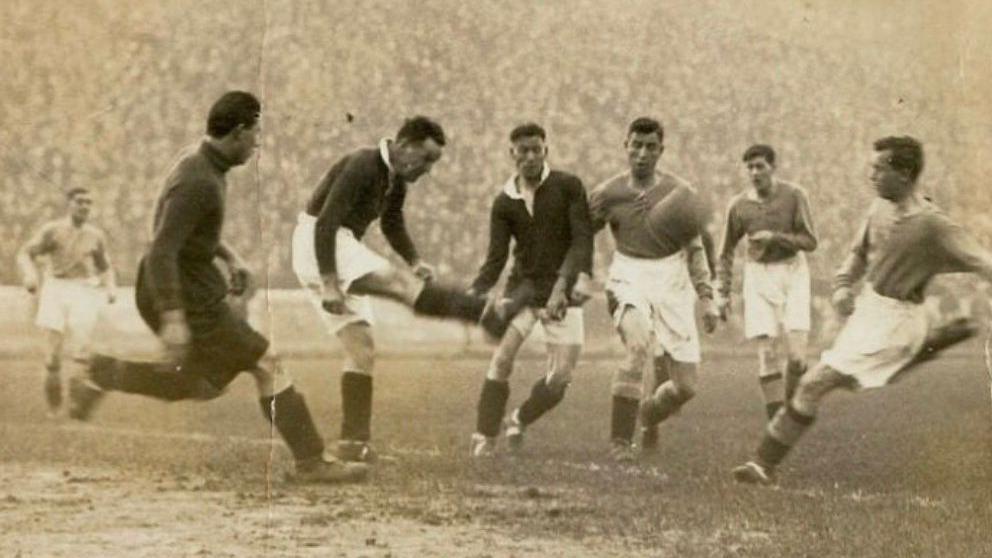 A black and white old photograph of a football match. Three players in black are competing for the ball with three other players in lighter-coloured kits.
