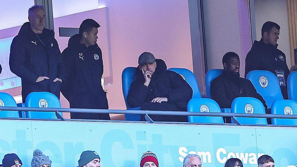 Pep Guardiola sits in front of a directors' box at Manchester City's Etihad Stadium