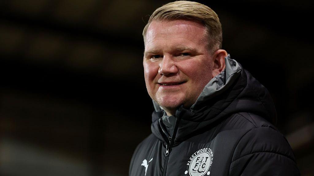 A smiling Pete Wild wearing a black coat with a white Fleetwood Town badge on the left breast