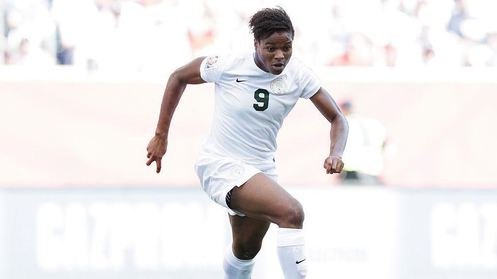 A Nigerian female player in an all-white shirt and short, with number 9 written on it with a green ink, in an action mood on a football field.