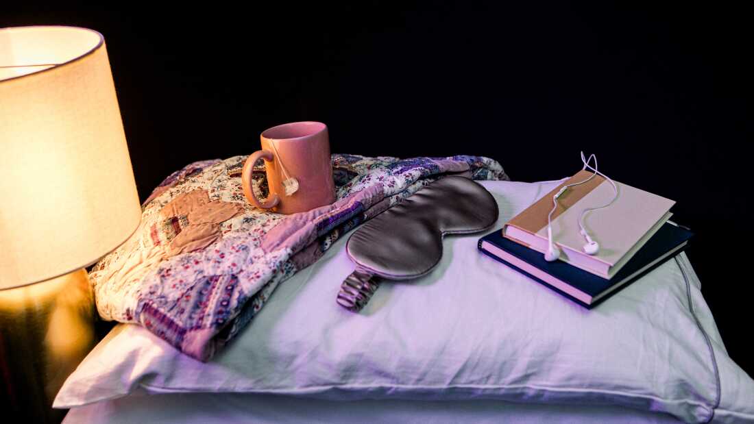 A pink cup of hot tea, a gray silk sleep eye mask, two books, and some wired headphones are laid out on top of two stacked pillows next to a table lamp that's turned on, emitting warm light. All the objects are photographed in a studio against a black backdrop.