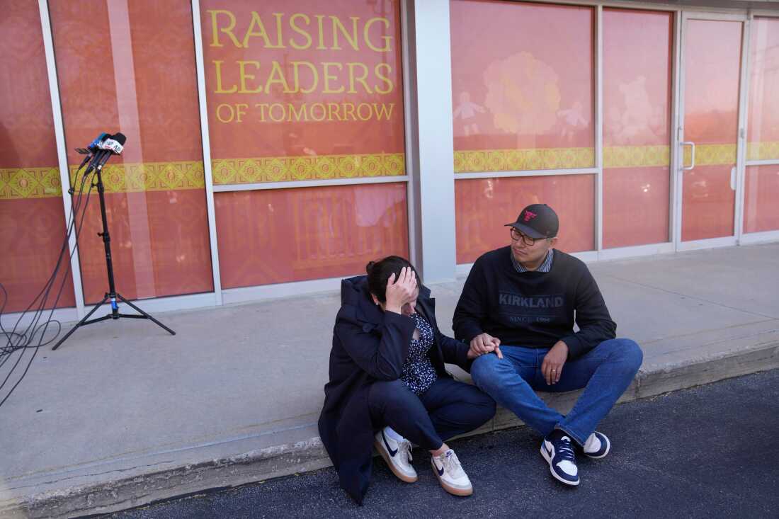 A man and a woman sit on a sidewalk curb in front of the school which is in a strip mall. The woman has her head in one hand and the man is holding her other hand. A sign behind them reads, "Raising Leaders of Tomorrow."