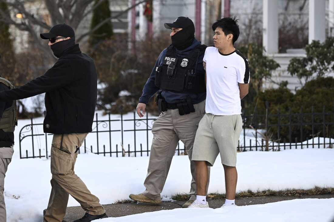 Masked ICE agents lead an Asian man in handcuffs. Snow is on the ground. The agents wear heavy winter gear; the man in custody is wearing short sleeves and shorts.